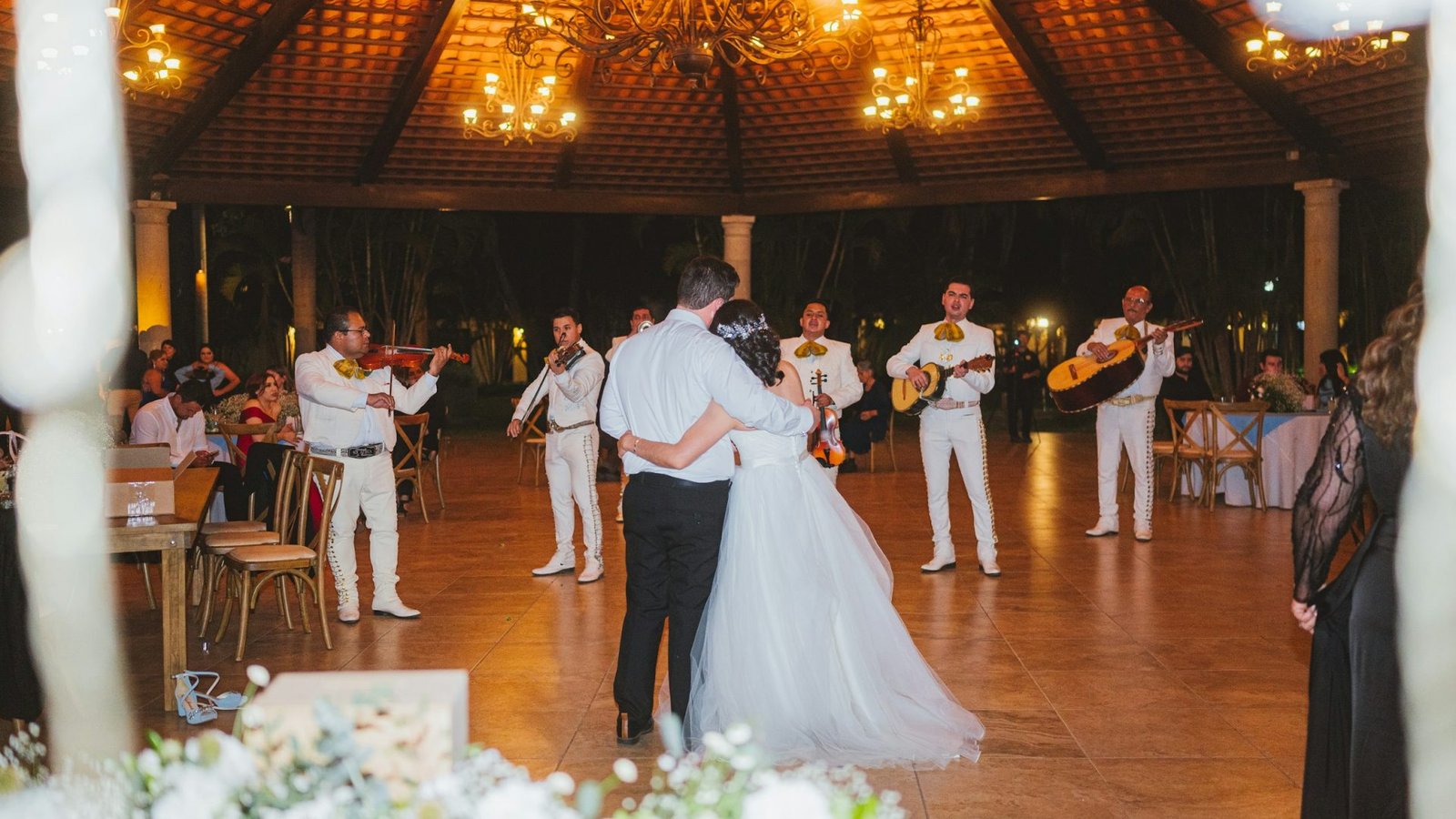 Trio de mariachi tocando ao vivo enquanto um casal de noivos dança durante a recepção do casamento, sob luz quente de lustres.